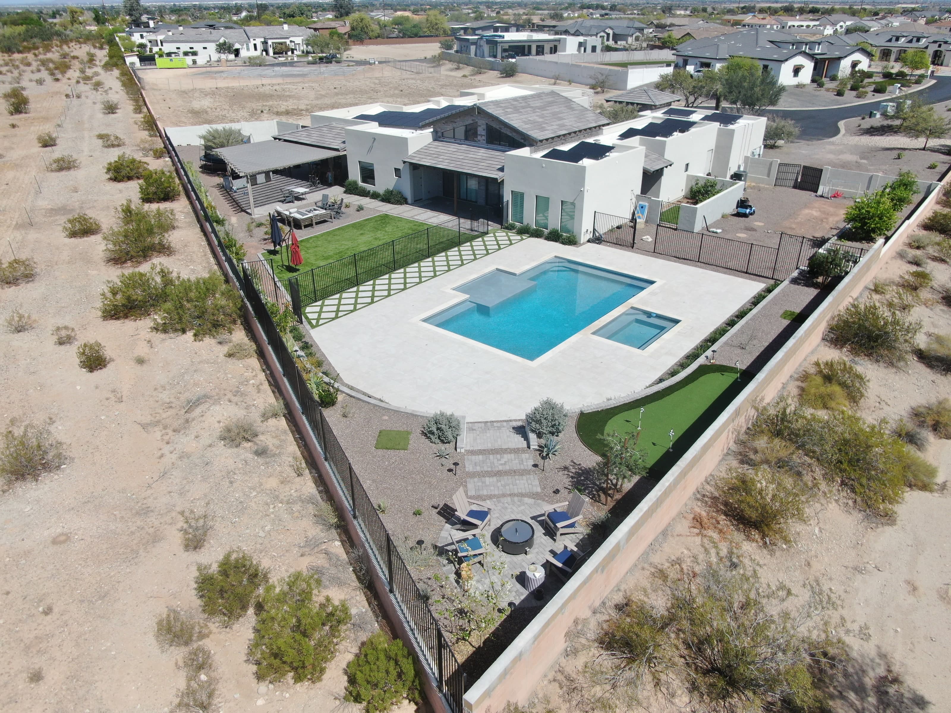 Aerial view of a finished backyard with a pool, spa, diamond turf-and-paver pattern, and putting green against a desert backdrop